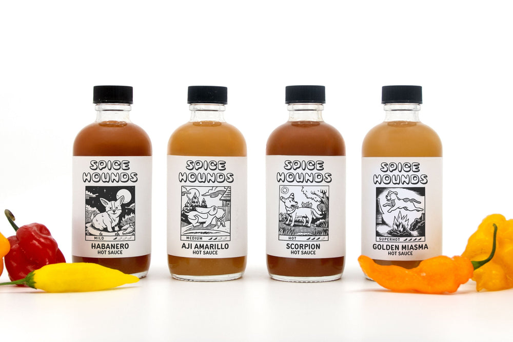 Four bottles of Spice Hounds hot sauce in a gift set & sampler called The Doghouse, displayed with hot peppers on a white background